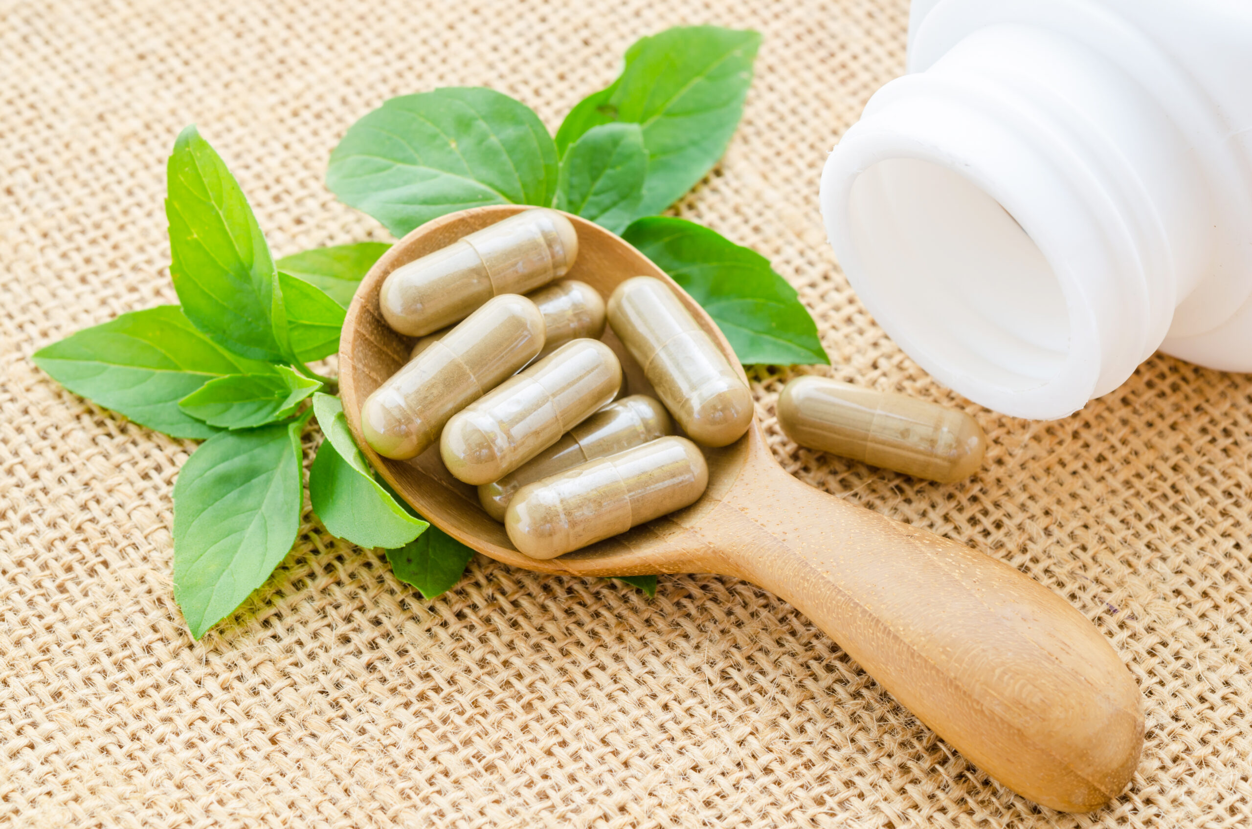 Herb capsule spilling out of a bottle in wooden spoon with green leaf on sack background.