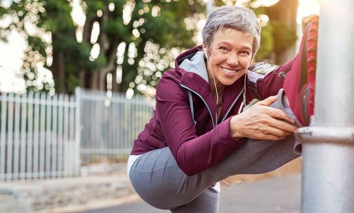Smiling retired woman listening to music while stretching legs outdoors. Senior woman enjoying daily routine warming up before running at morning. Sporty lady doing leg stretches before workout and looking at camera.